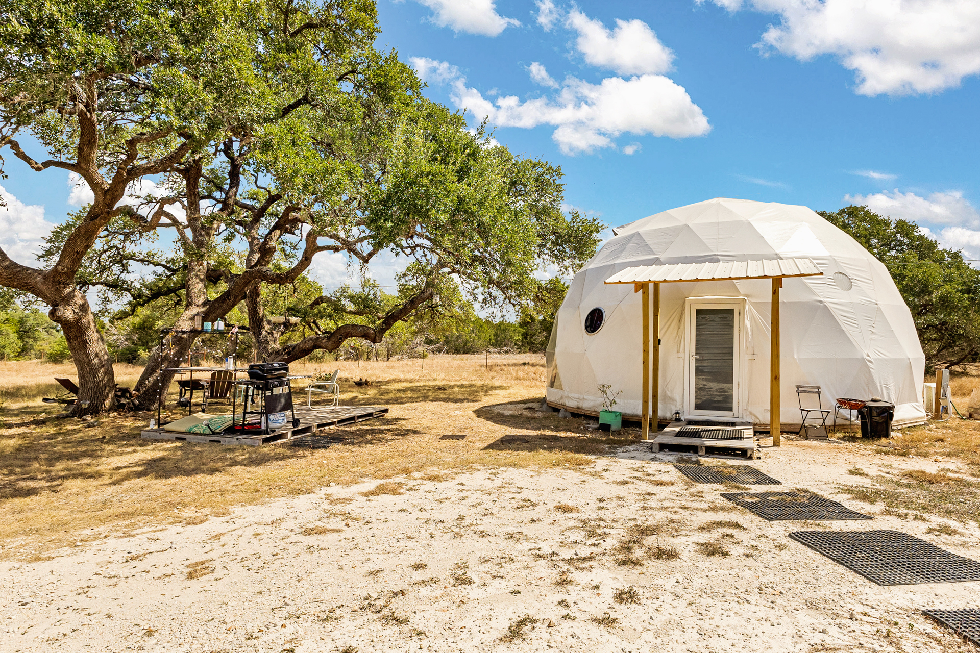 Glamping campsite featuring a geodesic dome, outdoor kitchen, and fire pit nestled under tall trees in the Texas Hill Country.
