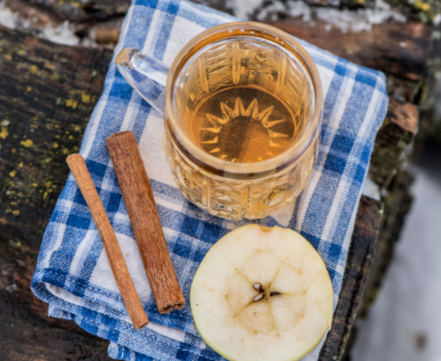 A glass mug of warm wassail sits on a blue-and-white checkered cloth beside two cinnamon sticks and a fresh apple slice.